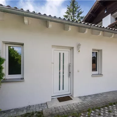 Ein modernes Haus mit einem weißen Eingang und großen Fenstern. Der Außenbereich ist mit Steinen gepflastert und von Grün umgeben.