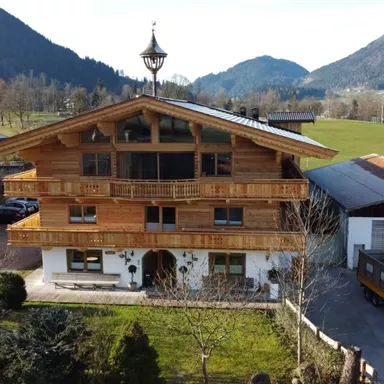 Ein rustikales Holzhaupthaus umgeben von grüner Landschaft und Bergen. Die Architektur zeigt alpine Elemente und eine einladende Veranda.
