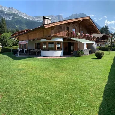 A charming wooden house with a large garden. In the background, majestic mountains and a blue sky can be seen.