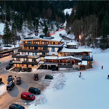 Ein modernes Gebäude im Schnee, umgeben von Wäldern. Fahrzeuge stehen am Parkplatz, und im Hintergrund sind Skifahrer zu sehen.