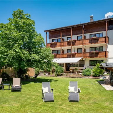 A building with several balconies and large windows surrounded by a well-maintained garden. In the foreground, there are lounges on a green meadow under a clear blue sky.