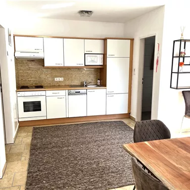 A modern kitchen with white furniture and a cooktop. The room is bright and inviting, with a large rug and a dining area.
