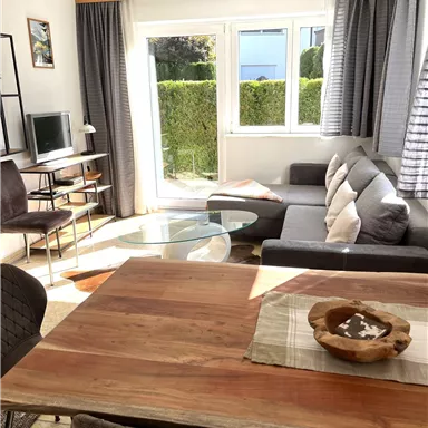 A modern living room with a cozy sofa and a wooden table.  
Large windows provide plenty of daylight and a view of the greenery.