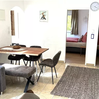 A bright living room with a dining table and cozy chairs. In the background, two beds and a window with a garden view can be seen.