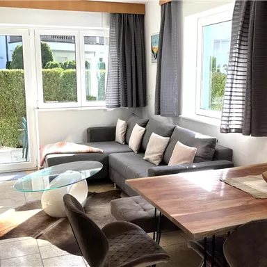 A bright and modern living kitchen with a cozy sofa and a dining table. Large windows let in plenty of daylight and offer a view of a well-maintained garden.