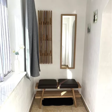 A bright entrance area with a wooden bench and a large mirror. A towel rack is hanging on the wall and a gray curtain is visible.