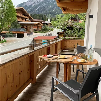 Ein gemütlicher Balkon mit Holzmöbeln und Blick auf die Berge. Auf dem Tisch stehen Snacks und Getränke.
