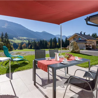 A cozy outdoor area with a table and chairs under a sunshade. In the background, mountains and a green landscape can be seen.