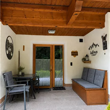 A cozy terrace with wooden beams and a large window. There is a table with chairs and a wooden bench.