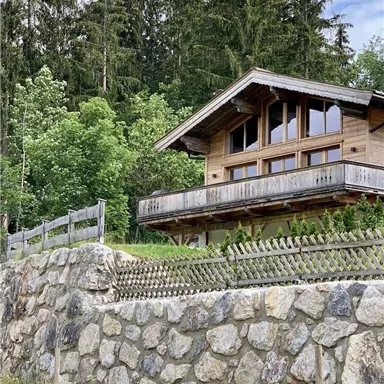 A charming wooden cottage with large windows, surrounded by trees. The garden is enclosed by a stone wall and a fence.