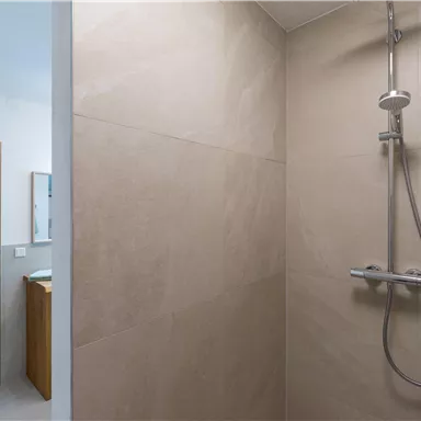 A modern shower with clear lines and bright tiles. In the background, there is a sink with a mirror visible.