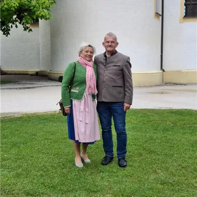 A couple is standing on a green meadow in front of a white building. The woman is wearing a traditional dress and a green jacket, while the man is wearing a gray jacket and jeans.