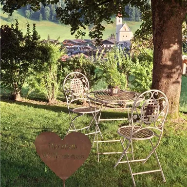 A cozy garden with a table and chairs under a tree. In the background, gentle hills and a small village can be seen.