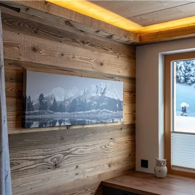 A cozy room with wood paneling and a large window. A landscape painting featuring snow-covered mountains hangs on the wall.