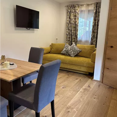 Ein gemütliches Wohnzimmer mit einem gelben Sofa und einem Esstisch. An der Wand hängt ein Fernseher und das Zimmer hat helle Holzfußböden.