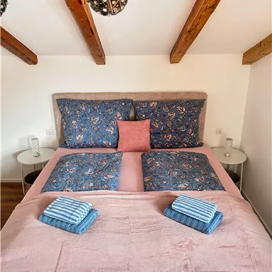 A cozy bedroom with a large bed dressed in blue bedding and adorned with a pink pillow. On the bed lie two striped towels, and the room features exposed wooden beams.