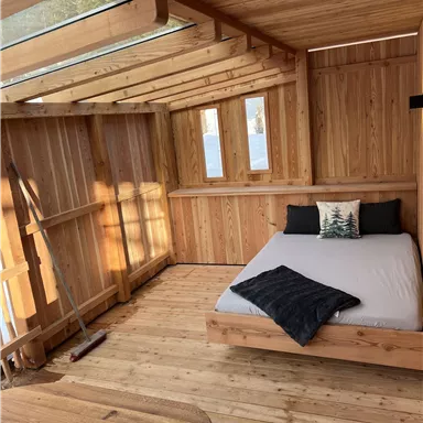 A bright wooden room with a double bed and large windows. The room has a cozy atmosphere and is simply furnished.