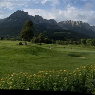 A vast golf course landscape with green grass and mountains in the background. The sky is partly cloudy, and the surroundings are peaceful and inviting.