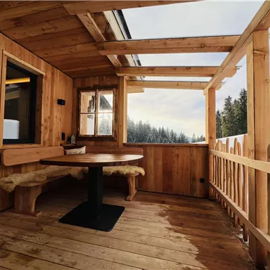 A cozy wooden terrace with a round table and seating. In the background, snow-covered trees can be seen.