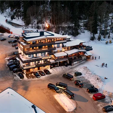 Ein modernes Gebäude im Schnee mit Autos auf einem Parkplatz. Im Hintergrund sind Skifahrer und eine schneebedeckte Landschaft zu sehen.