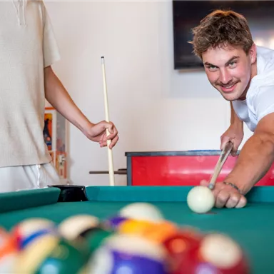 Two men are playing billiards in a bright room. One is smiling while he prepares a shot.