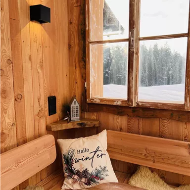 A cozy wooden cabin with a window overlooking snowy trees. On the bench lies a cushion with the inscription "Hello Winter."