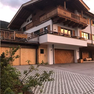 Ein modernes Haus im alpinen Stil mit einem Balkon und Holzverkleidungen. Der Eingang ist von einem gepflasterten Weg und grünen Pflanzen umgeben.