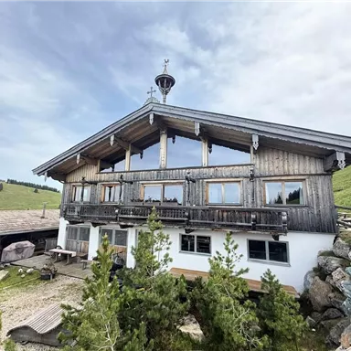 Eine traditionelle Berghütte aus Holz mit großen Fenstern. Die Umgebung ist grün und hügelig, ideal für einen ruhigen Rückzugsort.