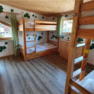 A cozy dormitory with wooden bunk beds. The walls are decorated with clovers, and large windows let in plenty of daylight.