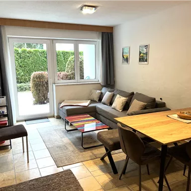 A modern living room with a gray couch and a dining table. Bright daylight shines through large windows, and the view overlooks a green garden.
