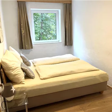 A cozy bedroom with a large bed and bright curtains. The view from the window shows green foliage.