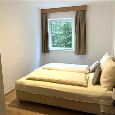 A cozy bedroom with a double bed and fresh bedding. The room has a window overlooking trees and light-colored walls.