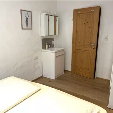 A simple bedroom with a double bed and a wooden wardrobe. The walls are white, and a wooden door leads outside.