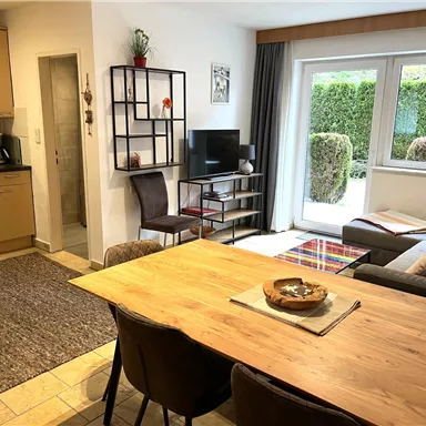 A bright living kitchen with a modern table and chairs. In the background is a cozy sofa and large windows that offer a view of a well-maintained garden.