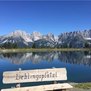 Eine idyllische Berglandschaft mit klaren Wasserflächen und einem Blick auf majestätische Gipfel. Im Vordergrund steht eine Bank mit der Aufschrift „Lieblingsplatzl“.