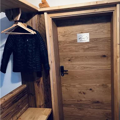 A cozy entrance area with a wooden bench, a hanging coat, and a wooden door. The room has rustic elements and a warm atmosphere.