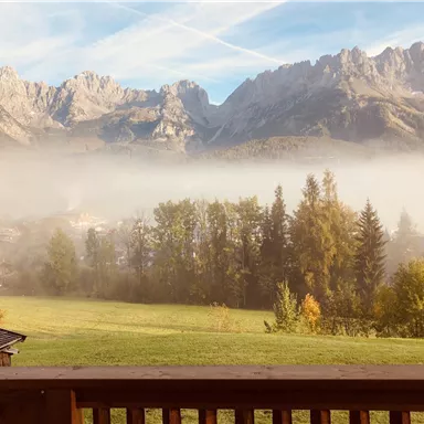 Eine wunderschöne Berglandschaft mit hohen Gipfeln und dichter Nebel. Im Vordergrund sind grüne Wiesen und Bäume zu sehen.