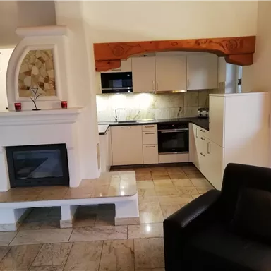 Eine moderne Küche mit weißen Schränken und einem eleganten Kamin. Neben der Küche steht ein schwarzes Sofa in einem hellen Raum.