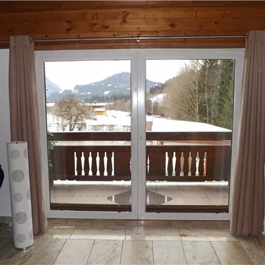 Ein geräumiges Zimmer mit großen Fenstern und Blick auf eine schneebedeckte Landschaft. Die Umgebung ist von Bergen und Bäumen geprägt.