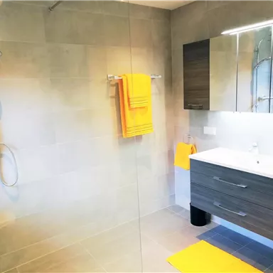 Ein modernes Badezimmer mit einer begehbaren Dusche und einem Waschbecken. Gelbe Handtücher und Matten sorgen für einen freundlichen Akzent.