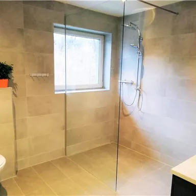Ein modernes Badezimmer mit einer Dusche aus Glas und einem weißen WC. Es gibt ein Fenster und einen kleinen Pflanzenbehälter auf der Fensterbank.