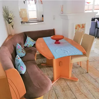 Ein gemütlicher Essbereich mit einer braunen Eckbank und einem Tisch. Auf dem Tisch steht eine rote Schale und bunte Kissen schmücken die Bank.