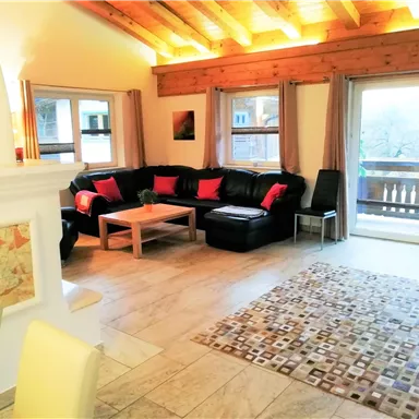 Ein gemütliches Wohnzimmer mit einer schwarzen Couch und bunten Kissen. Große Fenster bieten einen Blick auf einen Balkon und lassen viel Licht herein.