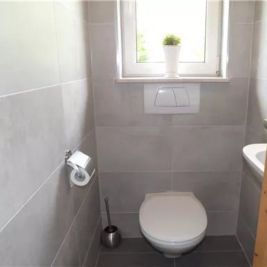 Ein kleines, modernes Badezimmer mit grauen Fliesen. Es ist mit einer Toilettenanlage und einem Fenster ausgestattet, das eine Pflanze zeigt.