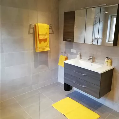 Ein modernes Badezimmer mit grauen Fliesen und einem schlichten Waschbecken. Gelbe Handtücher und eine Matte bringen einen farblichen Akzent in den Raum.