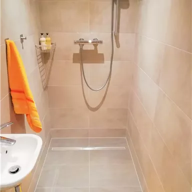 Ein modernes Duschbad mit grauen Fliesen und einer Handbrause. Ein orangefarbener Handtuch und ein gelber Badematten liegen bereit.