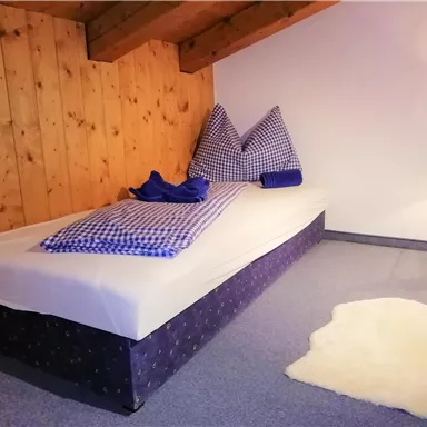 Ein einfaches Schlafzimmer mit einem Einzelbett und rustikalen Holzwänden. Auf dem Bett liegt eine blaue Decke und Kissen in kariertem Design.