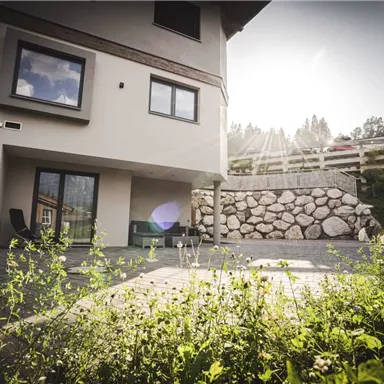 Ein modernes Haus mit einem sonnigen Außenbereich und einem geschmackvoll gestalteten Garten. Die Wände sind hell und die Fenster bieten einen Blick in die Umgebung.