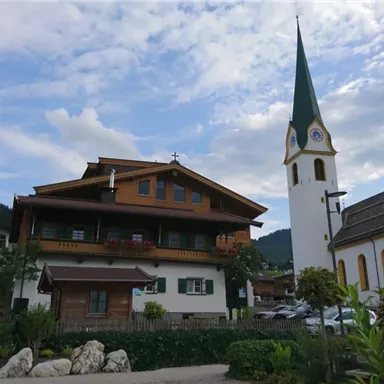 Ein malerisches Dorf mit einer traditionellen Kirche und einem Holzhaus. Der Himmel ist teilweise bewölkt und die Umgebung ist grün und einladend.