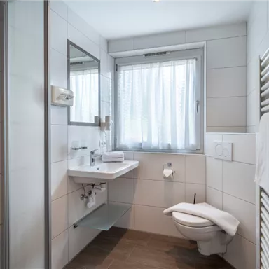 A modern bathroom with white tiles and a large window. It features a shower, a sink, and a toilet.